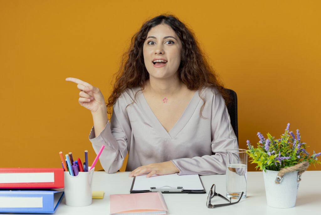 impressed young pretty female office worker sitting at desk with office tools points at side isolated on orange background with copy space